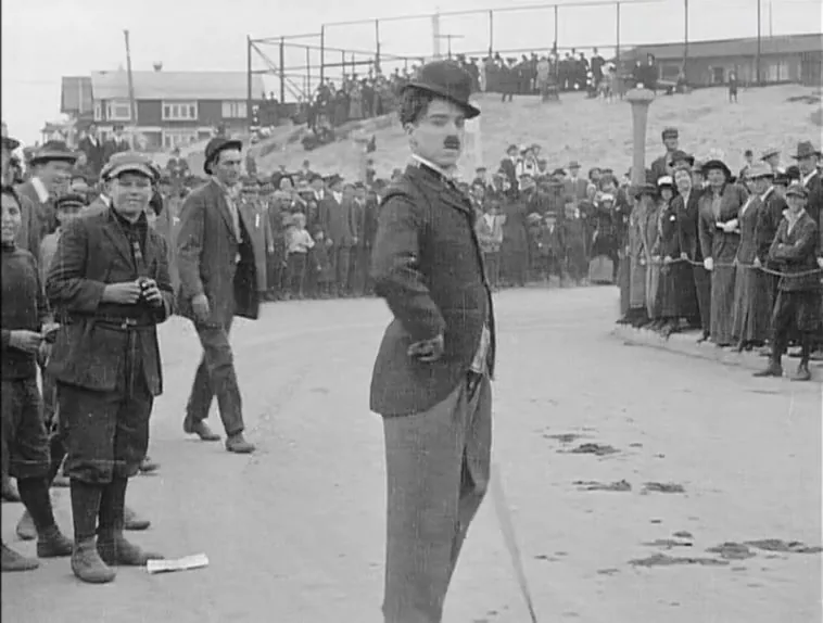 Бродяга стоит руки в бока у поворота трассы, за ним трибуна на холме, 1914