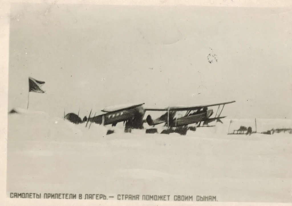 Два биплана Р-5 на лыжах на заснеженной льдине лагеря Шмидта, рядом красный флаг на шесте и нарты, апрель 1934 года