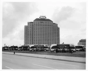 Futurliner с открытыми бортами-сценами стоят на пустыре перед высотным отелем Shamrock в Хьюстоне, у машин небольшие группы посетителей, февраль 1955 года