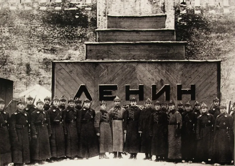 Курсанты в шинелях и будёновках стоят шеренгой перед первым деревянным Мавзолеем со ступенчатым верхом и надписью «Ленин», 1924 год
