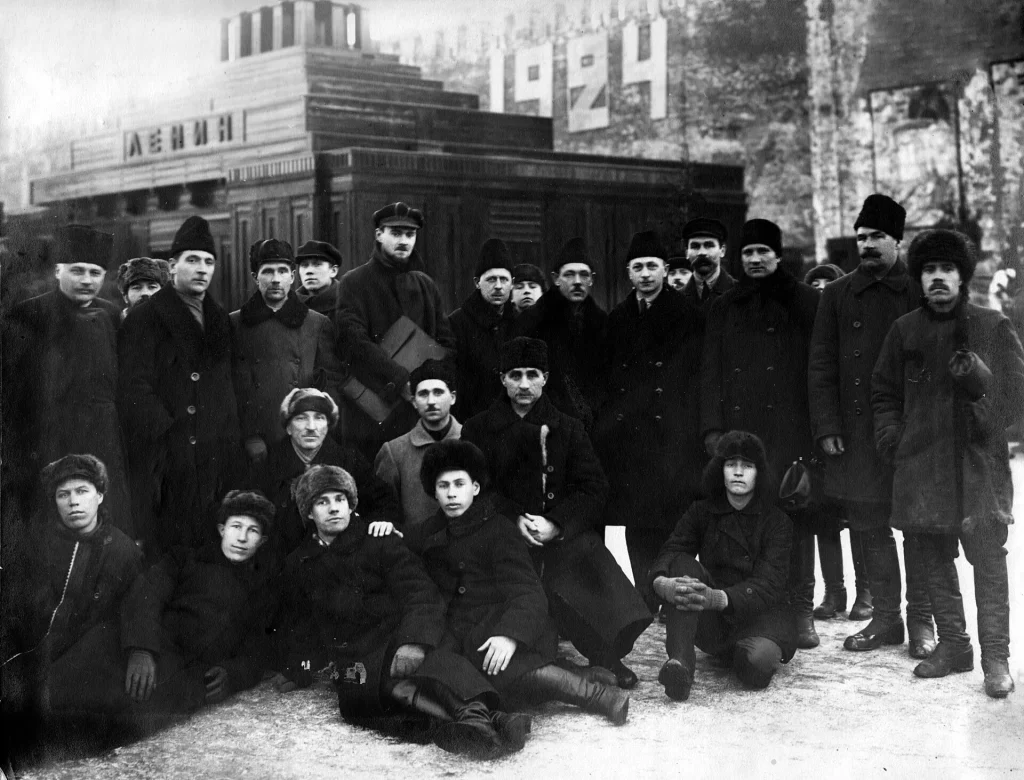 Группа мужчин в зимних шапках у второго деревянного Мавзолея с надписью «Ленин», на Кремлёвской стене крупные цифры «1924»