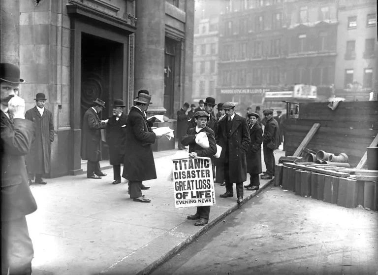 Мальчик-газетчик в кепке держит плакат «Titanic disaster great loss of life — Evening News» у входа в контору White Star Line в Лондоне, рядом мужчины в котелках читают газеты